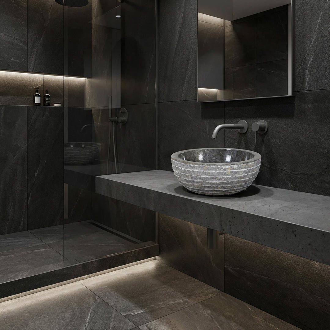 Round stone countertop basin on floating console, modern bathroom with dark tiles, glass shower and indirect light
