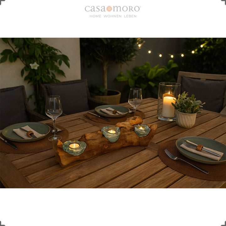 Three-part tea light holder made of glass and root wood as an atmospheric table decoration on a terrace, framed by plants and a set table